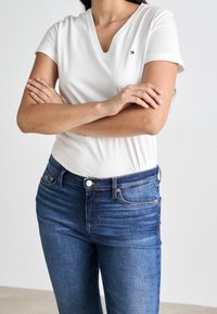 Camisa blanca de cuello en V hecha de tela suave, combinada con jeans de mezclilla azul oscuro con cierre de botón y diseño tradicional de cinco bolsillos.