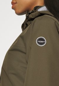 Profile of a person wearing an olive green jacket with a round black and white Icepeak logo patch on the upper sleeve.