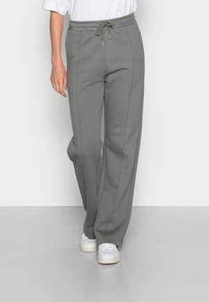 Personne portant un pantalon de survêtement gris avec un cordon de serrage, associé à des baskets blanches et une chemise blanche, debout devant un fond uni.