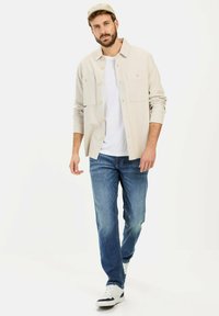 Beżowa koszula zapinana na guziki z dwoma kieszeniami na piersi, noszona na białą koszulkę, w połączeniu z wyblakłymi niebieskimi dżinsami i białymi trampkami, na jednolitym tle.