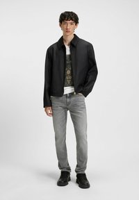 Sort, letvægtsjakke med krave, båret over en mønstret t-shirt. Kombineret med grå jeans og sorte sneakers. Ren design og pasform.