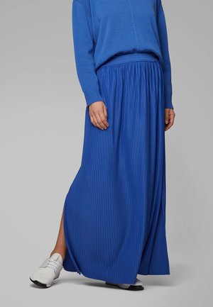 Woman wearing a bright blue pleated maxi skirt paired with a matching blue sweater and white sneakers on a gray background.