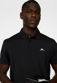 Svart polo skjorta med en texturerad krage och treknappslå; har en vit logotyp på vänster bröst. Modellen bär en svart handske.