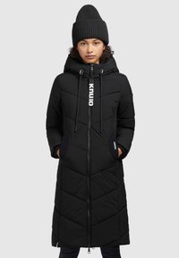Zwarte lange pufferjas met een capuchon, ritssluiting en zijzakken. Heeft een gewatteerd patroon en een logotag aan de voorkant. Vergezeld van een geribde zwarte beanie.