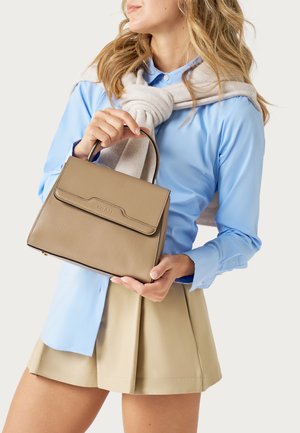 Vrouw in een blauw hemd en een beige rok die een taupekleurige leren handtas vasthoudt, met een lichtgrijze trui over haar schouders gedrapeerd.