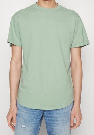 Homme portant un t-shirt à manches courtes vert clair uni et un jean bleu clair usé, debout devant un fond neutre.