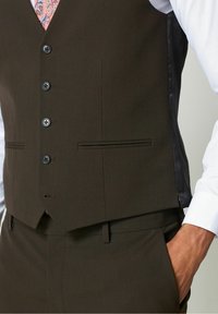 Eine Nahaufnahme einer braunen Weste eines Mannes mit schwarzen Knöpfen und seitlichen Einsätzen, die über einem weißen Hemd mit einem rosa gemusterten Krawatte getragen wird.