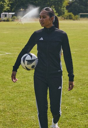 Sort atletisk træningsjakke og bukser med tre hvide striber, person som holder en sort-hvid fodbold på et græsareal.