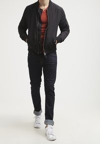 Mężczyzna idący, ubrany w czarną kurtkę typu bomber, czerwoną koszulę, ciemne jeansy ze zwiniętymi nogawkami oraz białe trampki na jednolitym białym tle.