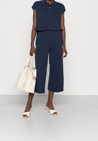 Navy blue collared jumpsuit with short sleeves and elastic waistband. White handbag with gold accents. Open-toe white heeled mules.