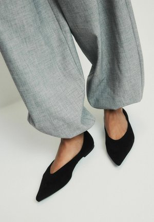 Legs wearing loose gray pants and black pointed-toe flats on a light background.