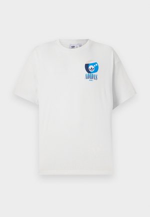 Mann trägt weißes Adidas-T-Shirt mit blauem Logo auf der Brust und schwarze Hose, zeigt Tätowierung am linken Unterarm vor einfachem Hintergrund.