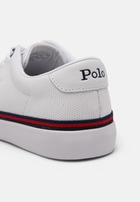 Baskets blanches perforées avec une bande rouge et marine sur la semelle et "Polo" brodé en noir sur la languette du talon.