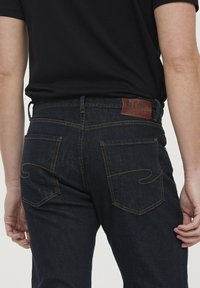 Jean en denim bleu foncé avec une étiquette en cuir portant le nom "Lee Cooper", des coutures jaunes et un design de poche arrière décontracté.