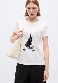 Άσπρο βαμβακερό T-shirt με γραφικό σχεδιασμένο σχήμα μιας φιγούρας με μαύρο παλτό και φούστα με πουά, κρατημένο από μοντέλο με κρεμ τσάντα.