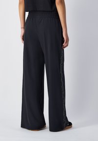Zwarte wijde broek met een smocktail in de taille, voorzien van een natuurlijk zijpatroon. Zacht, lichtgewicht materiaal met een vloeiende val.
