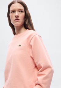 Γυναίκα με μακριά καστανά μαλλιά που φοράει ένα ανοιχτό ροζ φούτερ με μικρό πράσινο λογότυπο κροκοδείλου στο στήθος, κοιτάζοντας προς τα πλάγια.