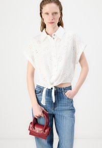 Chemise blanche à manches courtes avec un motif de broderie anglaise et un nœud sur le devant ; associée à un jean bleu à rayures taille haute. Tient un petit sac à main rouge.