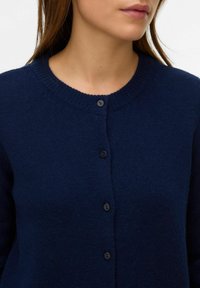 Námořnicky modrý pletený cardigan s kulatým výstřihem, šesti černými knoflíky a měkkou texturou, představující volný střih.