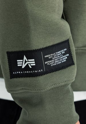Olivegrön sweatshirt med en svart etikett som har vit text och en logotyp. Tyget är slätt med ribbade muddarna och fållens detaljer.