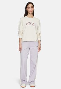 Crème kleurige sweatshirt met lange mouwen en een rood en blauw "FILA" logo. Lichtpaarse broek met witte zijpanelen en "FILA" print op de dij. Witte sneakers.