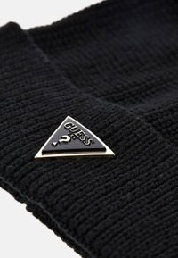 Berretto lavorato a maglia nero con texture a coste, dotato di una patacca triangolare oro e nera. Il materiale sembra morbido e caldo.