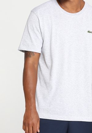 Hombre con camiseta de manga corta color gris claro con pequeño logo de cocodrilo verde en el lado izquierdo del pecho, pantalones azul marino y tatuaje visible en el brazo.