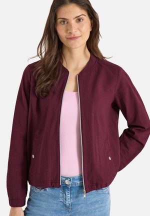 Kvinde iført en vinrød lynlåstrøje over en lyserød top og blå jeans, stående med den ene hånd på hoften mod en ensfarvet baggrund.