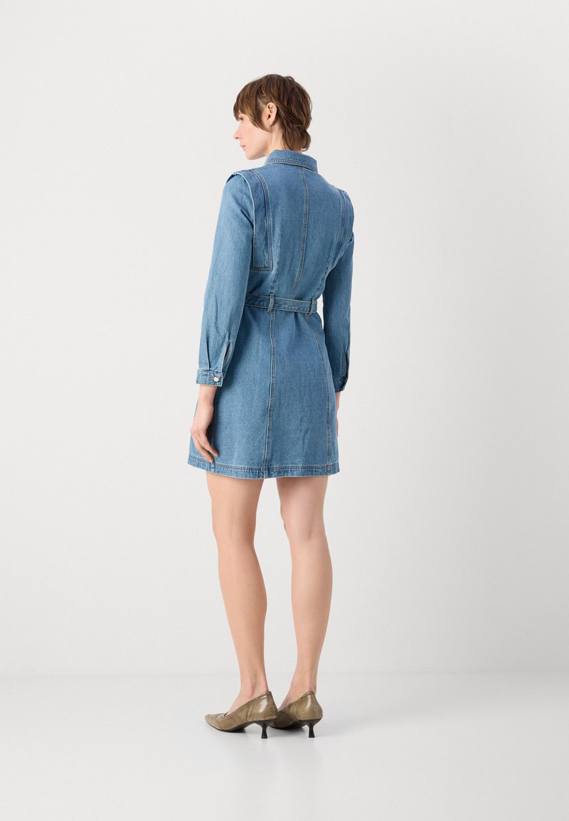 Femme debout, de dos, portant une robe en jean ceinturée à manches longues et des chaussures beiges à talons bas, sur un fond clair uni.