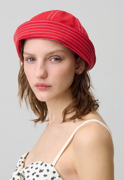 Cappello a secchiello rosso con dettagli di cucitura bianca, realizzato in tessuto. Il modello indossa un top a fantasia con spalline sottili e ha i capelli ricci.