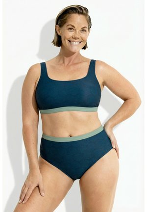 Glimlachende vrouw met kort haar draagt een donkergroene bikini met lichtgroene afwerking en patroon, poserend met één hand op haar heup tegen een effen achtergrond.