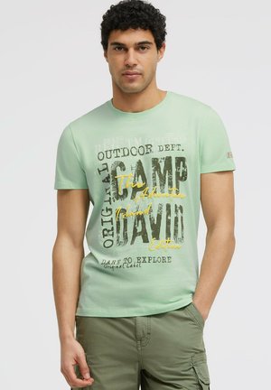 Mann mit lockigem Haar, der ein hellgrünes T-Shirt mit Grafik und olivfarbene Cargoshorts trägt, steht mit einer Hand in der Tasche.