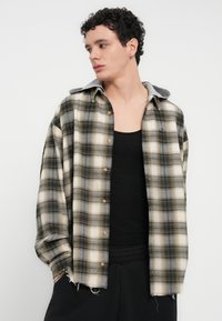 Καρό φανελένιο πουκάμισο σε μπεζ, μαύρο και γκρι, με γκρι γιακά. Διαθέτει κλείσιμο με κουμπιά και ξεφτισμένο τελείωμα, φορεμένο πάνω από μαύρο αμάνικο.