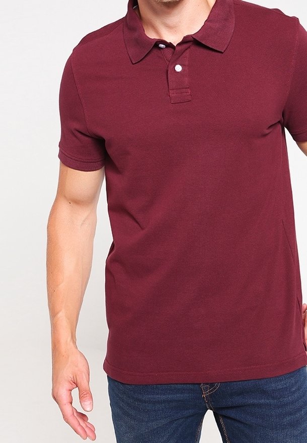 Homme portant un polo bordeaux à manches courtes et un jean bleu, debout devant un fond blanc uni.