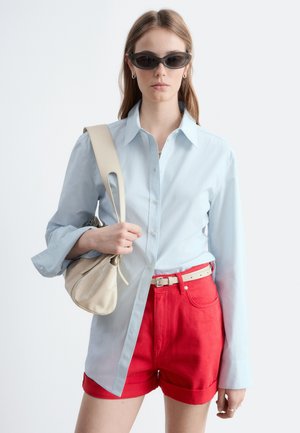 Ung kvinde iført sorte solbriller med katteøjne, lyseblå skjorte med knapper, røde shorts med højtalje, beige bælte og beige skuldertaske.