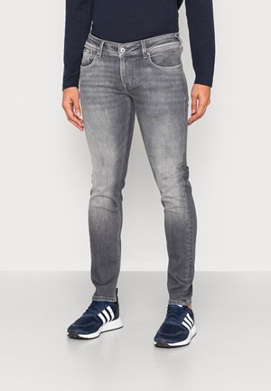 Mand iført slim-fit grå falmede jeans og navyblå sneakers med hvide striber, stående mod en ensfarvet baggrund.