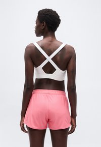 Γυναίκα, που φαίνεται από πίσω, φοράει λευκό αθλητικό σουτιέν με χιαστί πλάτη και ροζ σορτς Under Armour, μπροστά σε απλή φόντο.