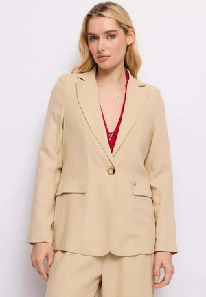 Donna bionda che indossa una giacca beige sopra una maglia rossa di pizzo, in piedi davanti a uno sfondo chiaro e uniforme.