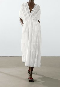 Personne à la peau foncée portant une robe midi blanche fluide et plissée avec des poches, ainsi que des sandales plates rouges et noires, debout sur un sol beige.