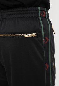 Lähikuva mustista pöksyistä, joissa on elastinen vyötärö, kultainen vetoketjutasku ja vihreä-punaiset raidat, joissa on tähti- ja nuolikuviot.