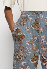 Lähekuva kädestä, joka on taskussa, päällä siniset kukkakuosiset housut, joissa on naru vyötäröllä, ja vaaleanbeige lyhythia shirt.