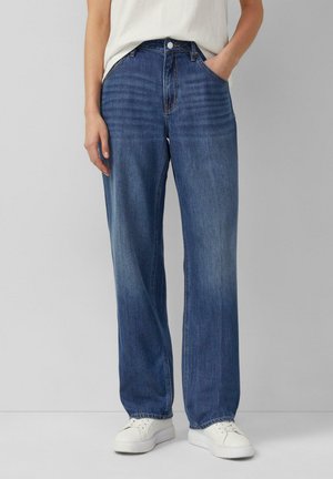 Osoba ubrana w niebieskie dżinsy z wysokim stanem i szerokimi nogawkami, połączone z białymi tenisówkami i włożonym do środka białym t-shirtem, stoi na tle szarego tła.