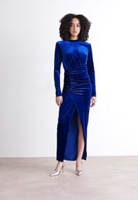 Vestido azul de veludo com mangas compridas, com franzido lateral e fenda alta na coxa. Usado com sapatos de salto alto prateados. Textura suave e design ajustado.