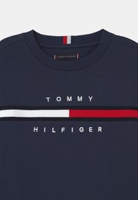 Sudadera azul marino con cuello redondo. Presenta una franja horizontal en bloques de color blanco, rojo y negro, con "TOMMY HILFIGER" en letras blancas.