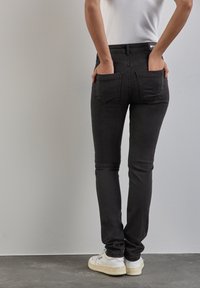 Svarta slim-fit jeans med en slät textur, två bakfickor och midjehöjd. Kombinerade med vita sneakers på ett grått golv.