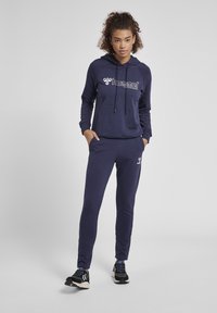 Marinblå hoodie med en framficka och dragsko, prydd med en vit logotyp. Matchas med matchande joggers, båda i ett mjukt tyg.