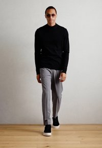 Camicetta nera a maniche lunghe, pantaloni grigi su misura, sneakers nere con suola bianca e occhiali da sole chiari, in piedi su un pavimento di legno.