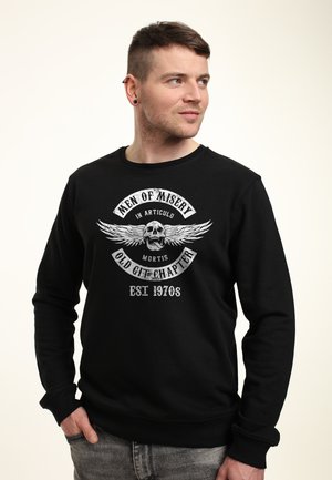 Czarny sweter z grafiką na przodzie, która przedstawia biały tekst i czaszkę ze skrzydłami. Okrągły dekolt i długie rękawy. Modna tekstura materiału.
