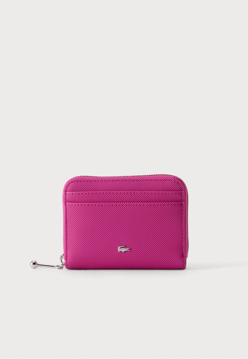 Portefeuille zippé rose vif avec surface texturée et petit emblème crocodile argenté à l'avant, posé sur un fond clair uni.