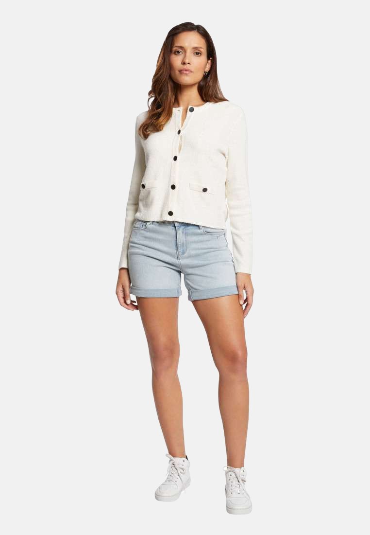 Cardigan blanc avec des boutons noirs, associé à un short en denim bleu clair et des baskets blanches. Le pull a une texture douce et un design décontracté.